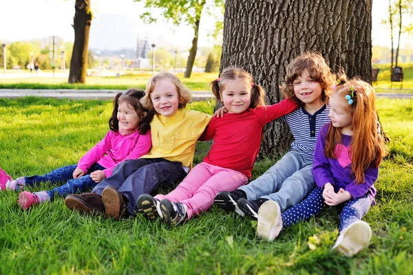 En grupp av små barn i färgglada kläder allomfattande sitter på gräset under ett träd i en park som skrattar och ler. — Stockfoto