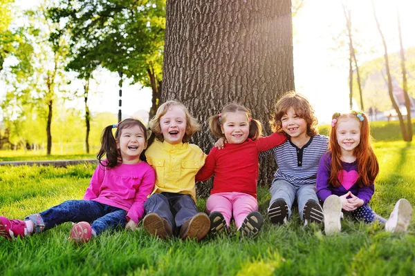 En grupp av små barn i färgglada kläder allomfattande sitter på gräset under ett träd i en park som skrattar och ler. — Stockfoto