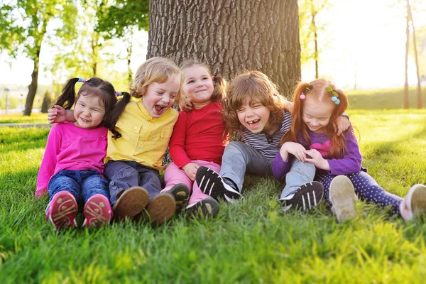 Un grupo de niños pequeños con ropa colorida abrazándose sentado en la hierba debajo de un árbol en un parque riendo y sonriendo . — Foto de Stock
