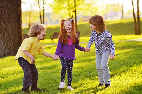 Un grupo de niños pequeños sonriendo tomados de la mano sobre un fondo de hierba, un árbol y un parque . —  Fotos de Stock