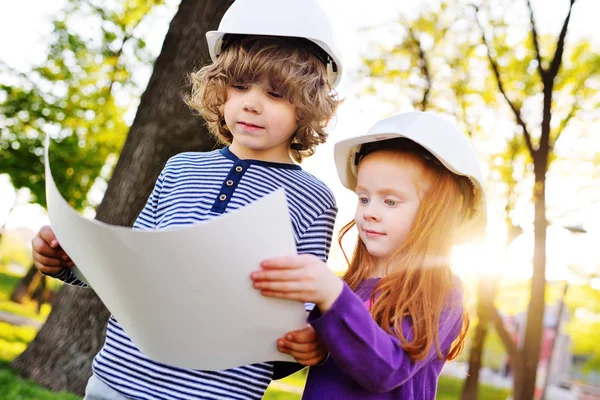 Anak laki-laki dan perempuan dalam helm konstruksi melihat lembaran putih kertas atau menggambar dan tersenyum — Stok Foto