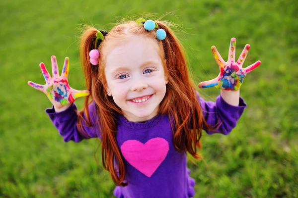 Little girl with red hair shows her hands dirty with multicolored paints and smiles — Stock Photo, Image