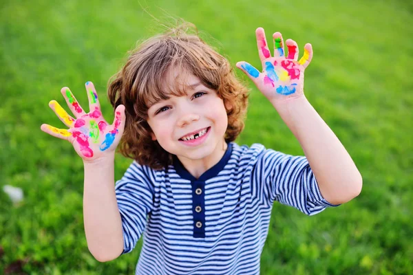 Jongen met krullend haar zonder melk voortand toont handen vuil met veelkleurige vinger verven en glimlacht. — Stockfoto