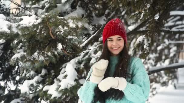 Een aantrekkelijke jonge vrouw in een warme gebreide blauwe trui en een rode hoed heeft plezier en danst tegen een achtergrond van besneeuwde bomen en sneeuw. — Stockvideo