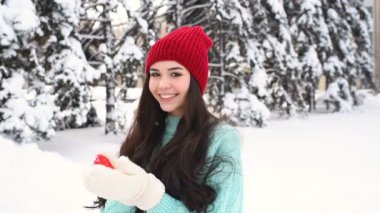Güzel genç kız sıcak mavi kazak ve kırmızı bir kalp tutan ve gülümseyen karla kaplı ağaçlar arka mittens