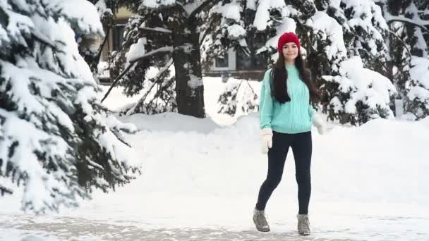 暖かいニットの青いセーターと赤い帽子の魅力的な若い女性は雪に覆われた木や雪を背景に楽しさとダンスをしています. — ストック動画