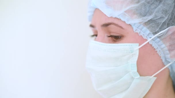 Jonge vrouwelijke arts of verpleegkundige met een medisch masker met droevige ogen op een witte achtergrond. Coronavirus, ziekte, covid-19, operatiekamer, chirurgie, verdriet. — Stockvideo