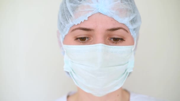 Ung kvinna läkare eller sjuksköterska i en medicinsk mask med ledsna ögon på en vit bakgrund. Coronavirus, sjukdom, covid-19, operationsrum, kirurgi, sorg. — Stockvideo