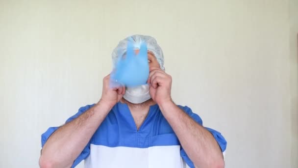 Komik doktor. Tıbbi maskeli bir adam beyaz arka planda lastik eldiven şişiriyor.. — Stok video