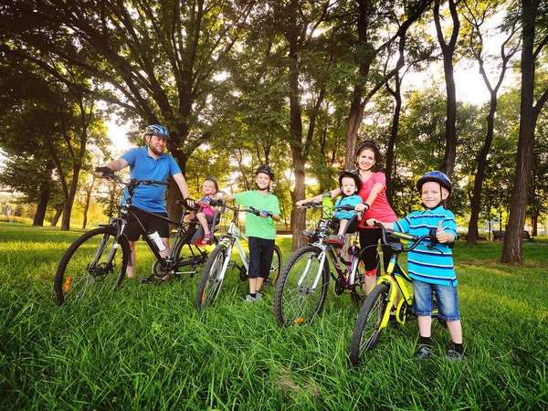 Grupo de personas en bicicletas: dos adultos y cuatro niños pequeños en equipo de bicicletas y cascos contra el fondo de los árboles, el parque y la hierba verde. Estilo de vida familiar y activo . —  Fotos de Stock
