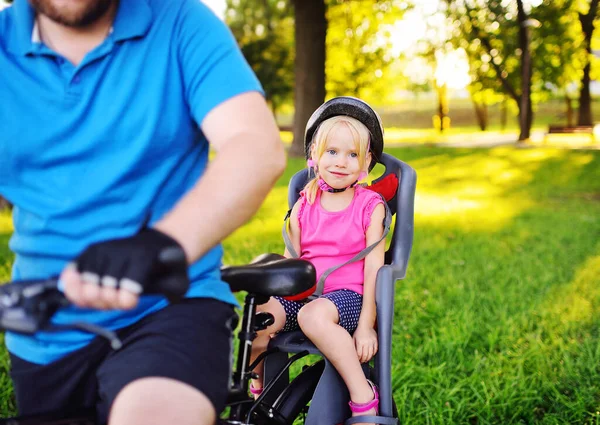 Niño una niña pequeña en un casco de bicicleta sonríe sentado en un asiento para niños bicicleta contra el fondo de un parque y hierba verde . — Foto de Stock