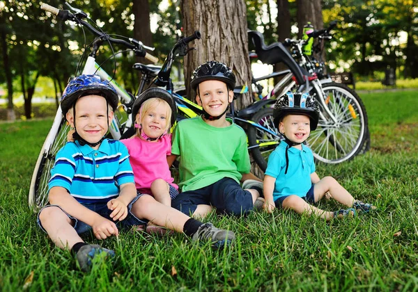 Grupo de niños pequeños en edad preescolar Cascos de seguridad de bicicletas sonríen sentados en la hierba verde fresca en el Parque contra el fondo de bicicletas y árboles . —  Fotos de Stock