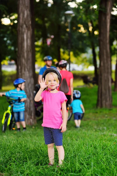 Una niña pequeña con una camiseta rosa y un casco de bicicleta protector sonríe y se agita contra el fondo de sus padres y familiares en bicicletas en el parque . — Foto de Stock