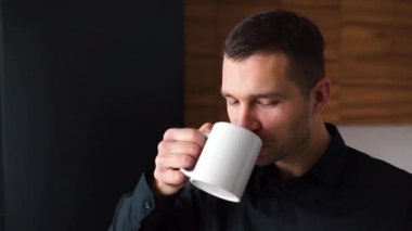 Siyah gömlekli, sıcak içecek içen ve tablet kullanan başarılı bir genç adam videosu. Evden uzak bir iş. Kamera birkaç kez yukarı ve aşağı hareket eder.