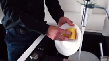 Siyah elbiseli adama yaklaş lavabonun önünde dur ve musluğu aç. Bulaşık yıkamak için su akıyor. Adam birbiri ardına beyaz tabakları temizliyor..