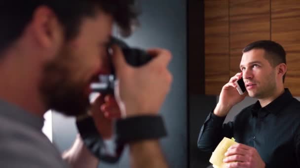 Proces van het nemen van foto 's of het opnemen van video. Jonge zakenman in zwart shirt aan de telefoon tijdens de lunch. Hou een boterham vast. Zakelijk gesprek. Werk op afstand van huis. — Stockvideo