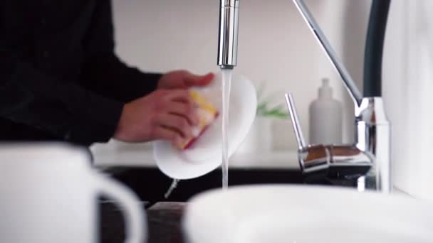 Διαδικασία καθαρισμού λευκού πιάτου με σφουγγάρι και χέρια. Ο τύπος στέκεται στο νεροχύτη και ανοίγει τη βρύση με νερό. Το κλείνω αφού τελειώσω το πλύσιμο των πιάτων. Πλευρική άποψη. — Αρχείο Βίντεο