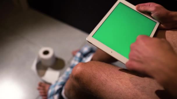 男人坐在马桶或壶上，手里拿着绿色屏幕的平板电脑的视频。跳下去好几次了地板上的卫生纸卷. — 图库视频影像