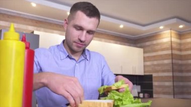 Sandviç yapma süreciyle ilgili video kaydı. Genç adam biraz ketçap ve hardal ekleyip üzerine peynir ve marul koy. Yemeğini bitir ve lezzetli bir şeyler yemeye hazır ol.