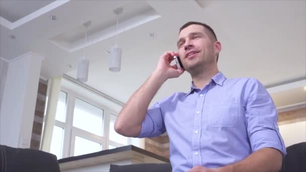 Low view of young man talking on smartphone and watching tv. Switching channels using remote control. Positive cheerful attractive guy smiling during conversation. — Stock Video