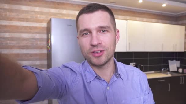 Jovem blogueiro do sexo masculino gravando vídeo em casa. Segure a câmera com a mão e fale. Ponto na mobília de cozinha traseira. Conversa alegre e confiante . — Vídeo de Stock