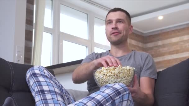 Boldog pozitív fiatalember pizsamában tévécsatornát vált távirányítóval és nevetett. Popcornt enni a szobában és élvezni. Bűn otthon a kanapén. — Stock videók