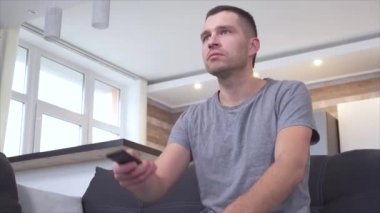 Ev elbiseli genç adam televizyon izlemeye ve uzaktan kumandayla açmaya hazır. Televizyon dizisi ya da film izliyordum. Hoşgörü ve şok içeriği. Adam kötü filme tepki veriyor..