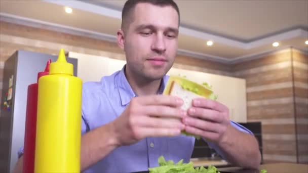 Video van de jongeman zit en eet smakelijke zelfgemaakte sandwich. Voeg wat sla met mosterd en ketchup toe. Vettig maar heerlijk eten. Nog meer kauwen en bijten. — Stockvideo