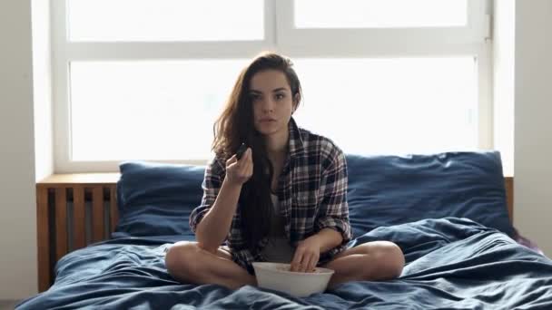 Una joven se queda en casa durante la cuarentena. Chica sentarse en la cama y cambiar los canales de televisión. Comiendo palomitas. Asustado un poco y sorprendido de ver la película solo en la habitación . — Vídeos de Stock