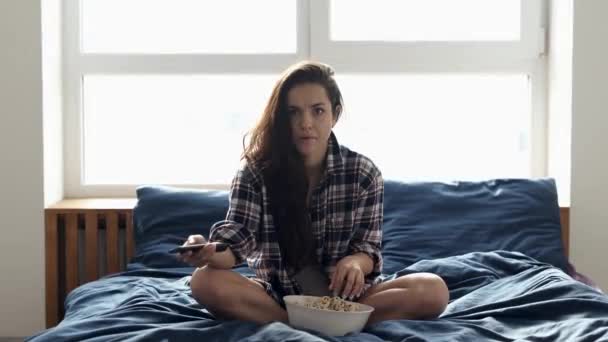 若い女性は隔離中に家にいます。女の子ベッドの上に座って、映画を見るのを恐れている。ポップコーンを食べてテレビチャンネルを切り替える. — ストック動画