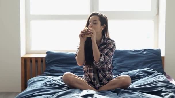 Una joven se queda en casa durante la cuarentena. Siéntese en la cama y comer delicioso sándwich de grasa sabrosa. Disfruta mordiendo pan fresco. Comida Divour sola en la habitación . — Vídeos de Stock