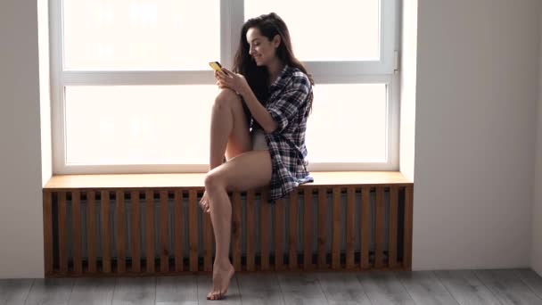 Ung kvinna stannar hemma under karantänen. Chatta online med hjälp av smartphone. Sitt vid fönstret i pyjamas och le. Smal välbyggd kvinna på morgonen. — Stockvideo