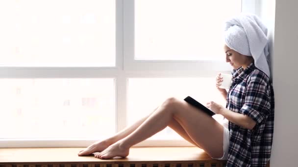 Молода жінка залишається вдома під час карантину. Сідайте у вікно після купання та читання книги. Пийте чай або каву і насолоджуйтеся читанням. Сміючись з бадьорістю . — стокове відео