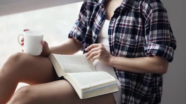 Jonge vrouw blijft thuis tijdens quarantaine. Ga aan het raam zitten met boek en kopje thee of koffie. Sla pagina 's om en stop ermee. Bovenaanzicht. — Stockvideo