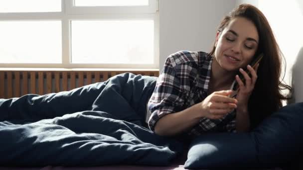 Νεαρή γυναίκα μένει σπίτι κατά τη διάρκεια της καραντίνας. Πληκτρολογώντας τον αριθμό τηλεφώνου στην οθόνη smartphone και μιλώντας με ευχαρίστηση. Ξαπλωμένος στο κρεβάτι το πρωί. Ρομαντική συζήτηση. Αργή κίνηση. — Αρχείο Βίντεο