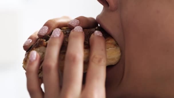 La jeune femme reste à la maison pendant la quarantaine. Vue latérale et gros plan de fille manger un pain ou un hamburger savoureux. Mordre gros morceau et mâcher. Isolé sur fond blanc. Mouvement lent . — Video