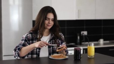 Karantina sırasında mutfakta genç bir kadın. Evde, masada kal ve ekmeğin üzerine biraz bıçaklı reçel koy. Lezzetli tatlı kahvaltı. Zevkle yiyorum..