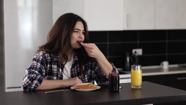 Jovem na cozinha durante a quarentena. Menina sentar-se à mesa e comer pão torrado com geléia. Desfrutando de café da manhã ou refeição matinal com suco de laranja . — Vídeo de Stock