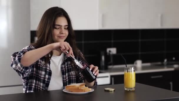 Giovane donna in cucina durante la quarantena. Metti un po 'di marmellata fuori dal barattolo sul pane tostato e ungilo. Colazione in mattinata. Godersi il dolce pane tostato . — Video Stock