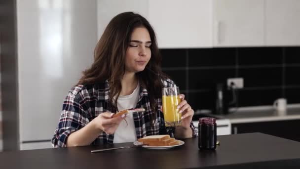 Νεαρή γυναίκα στην κουζίνα κατά τη διάρκεια καραντίνας. Κορίτσι που τρώει ψωμί με μαρμελάδα και χαμόγελο. Χαρούμενη γυναίκα που πίνει χυμό πορτοκάλι. Απολαμβάνοντας πρωινό το πρωί. — Αρχείο Βίντεο