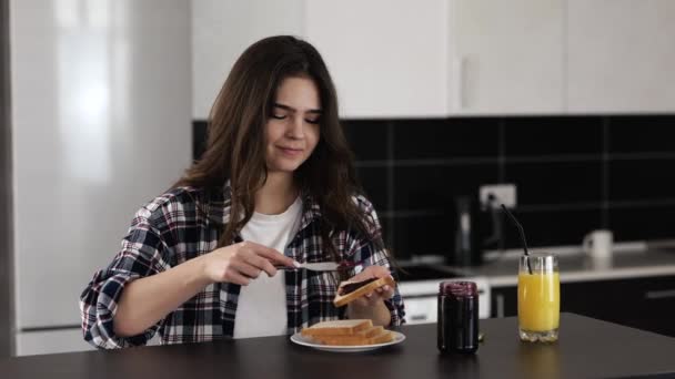 Νεαρή γυναίκα στην κουζίνα κατά τη διάρκεια καραντίνας. Μείνε σπίτι στο τραπέζι και βάλε μαρμελάδα με μαχαίρι στο ψωμί του τοστ. Υπέροχο γλυκό πρωινό. Φαγητό με ευχαρίστηση. — Αρχείο Βίντεο