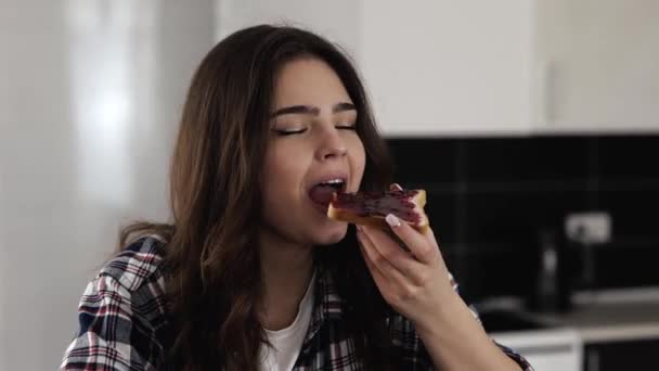 Mladá žena v kuchyni během karantény. Sedněte si a jezte chutné chutné kus chleba s džemem. Ráno si vychutnávám snídani. — Stock video
