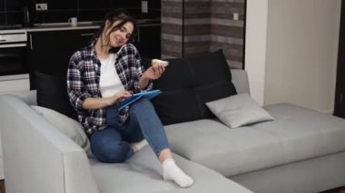 Karantina sırasında mutfakta genç bir kadın. Kanepeye otur ve tablet kullanarak internet sörfü yap. Telefonda konuşmak ve sandviç yemek. Ev kumandasında çalışmak.