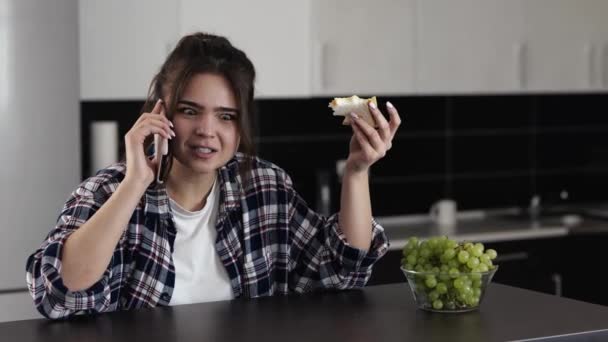 Mujer joven en la cocina durante la cuarentena. Disfrutando de comer sándwich con queso y conversación telefónica. Hablar emocionalmente en el smartphone . — Vídeos de Stock