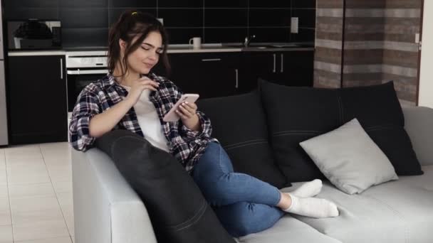 Mujer joven en la cocina durante la cuarentena. Chica sentarse en el sofá y escribir en el teléfono inteligente. Redes sociales en línea. Sonriendo durante el mensaje de escritura . — Vídeo de stock