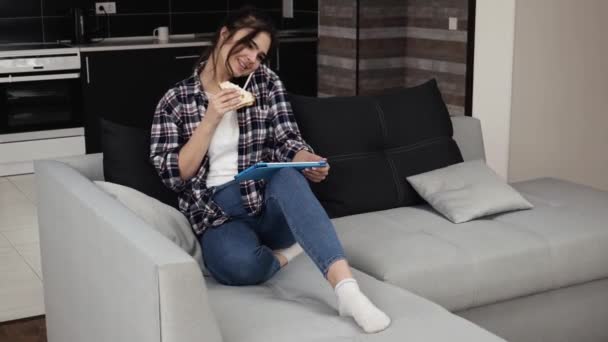 Mujer joven en la cocina durante la cuarentena. Hablar por teléfono y el uso de la tableta para navegar por Internet en línea. Siéntate en el sofá y come sándwich. Trabajo remoto . — Vídeo de stock