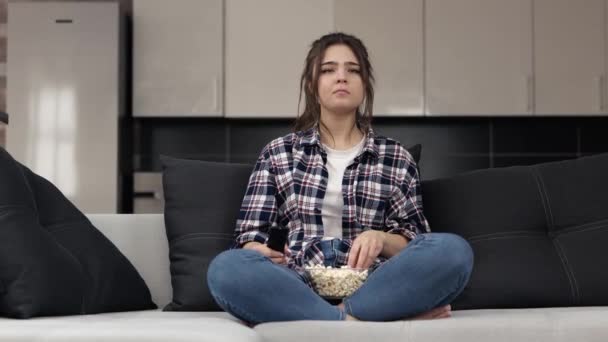 Mladá žena v kuchyni během karantény. Sedněte si na pohovku se zkříženýma rukama a dívejte se na film nebo televizi. Překvapený a šokovaný. — Stock video