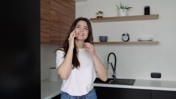 Giovane donna in cucina durante la quarantena. Alzati e parla sullo smartphone. Conversazione online. Bella ragazza sorridente e ridente . — Video Stock