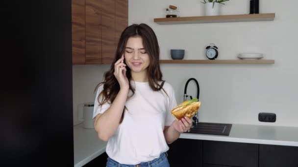 Mujer joven en la cocina durante la cuarentena. Hablando en el teléfono inteligente y sosteniendo sándwich en la mano. Expresión emocional. Hora de comer sándwich . — Vídeo de stock