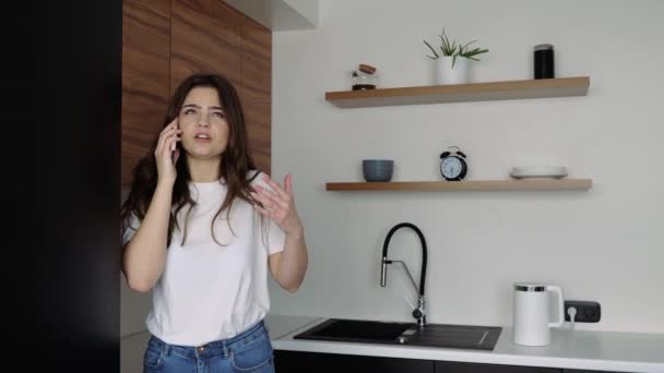 Mujer joven en la cocina durante la cuarentena. Hablando por teléfono emocionalmente. Grita y discute. Terminar de hablar por teléfono y deslizar la información en la pantalla . — Vídeo de stock